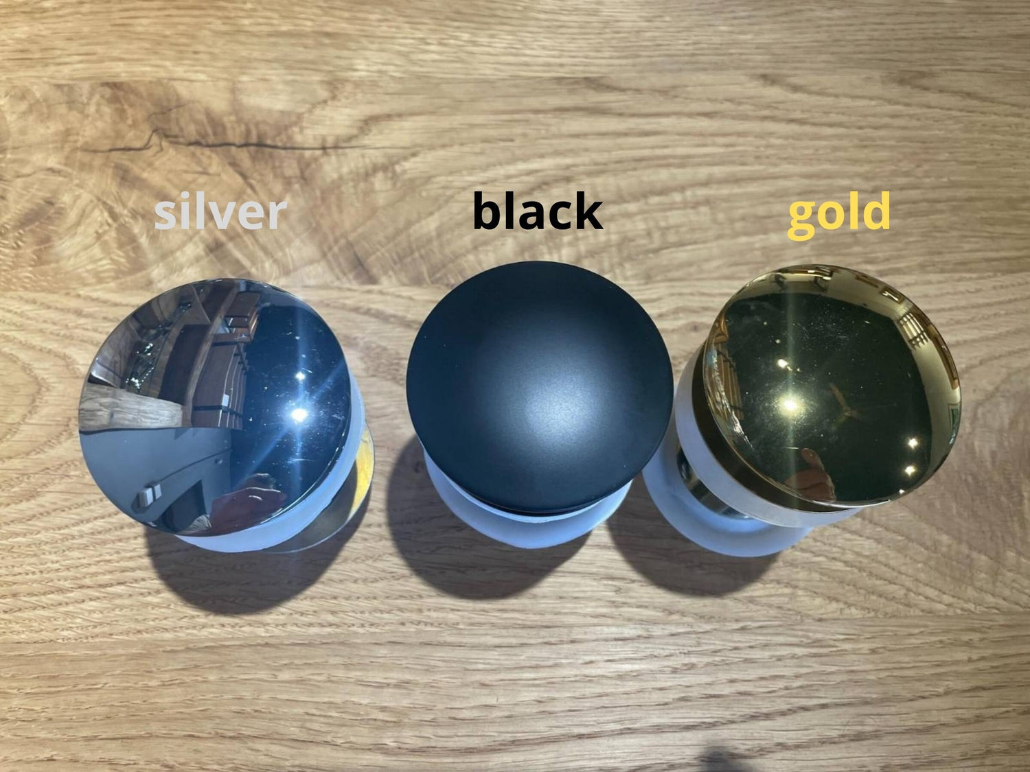 pop-up sink drain stoppers in silver, black, and gold finish displayed on oak wood surface – drain plugs for wooden washbasins
